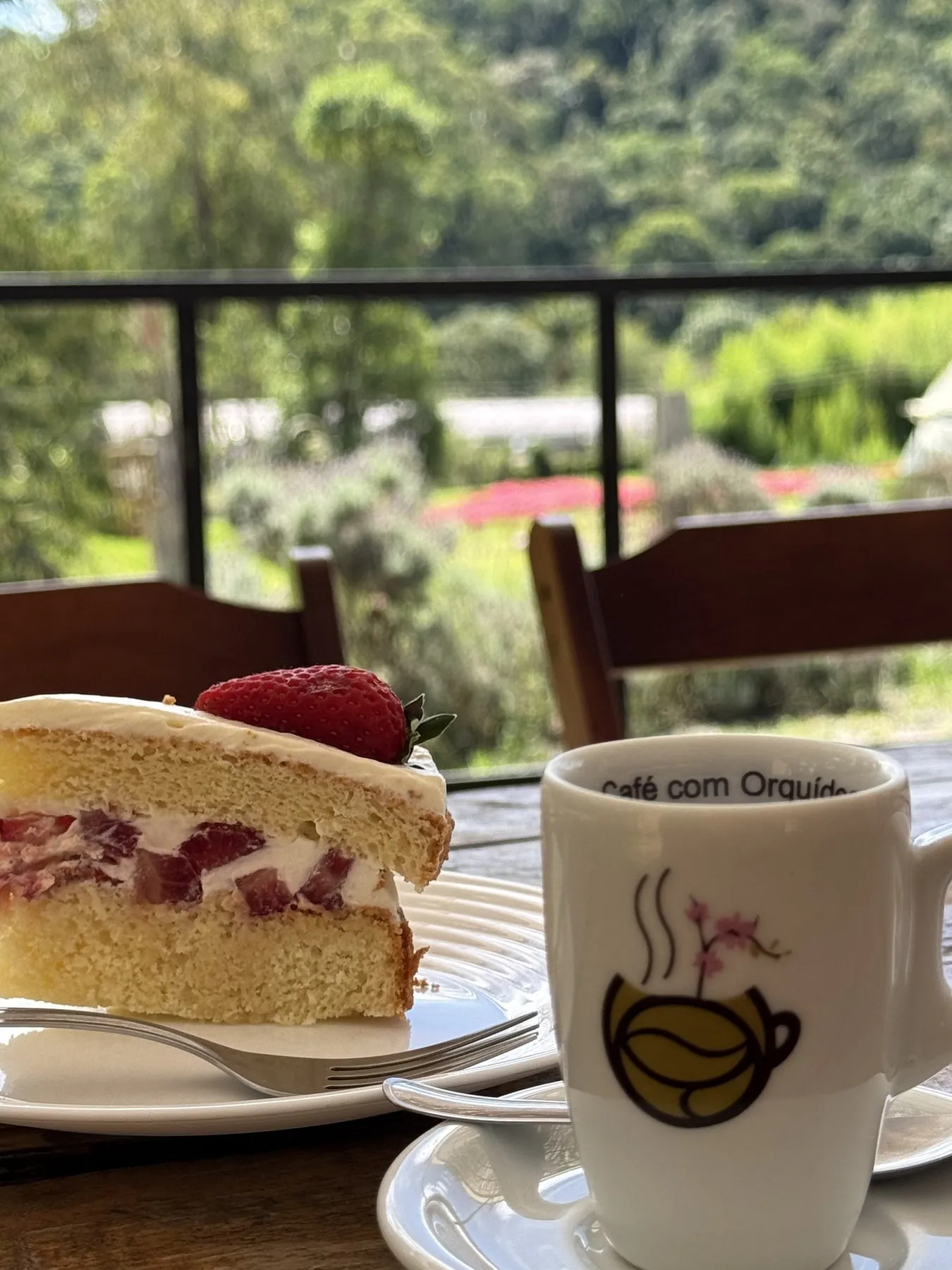 Fatia de bolo de morango e xícara do Café com Orquídea, com o jardim florido ao fundo